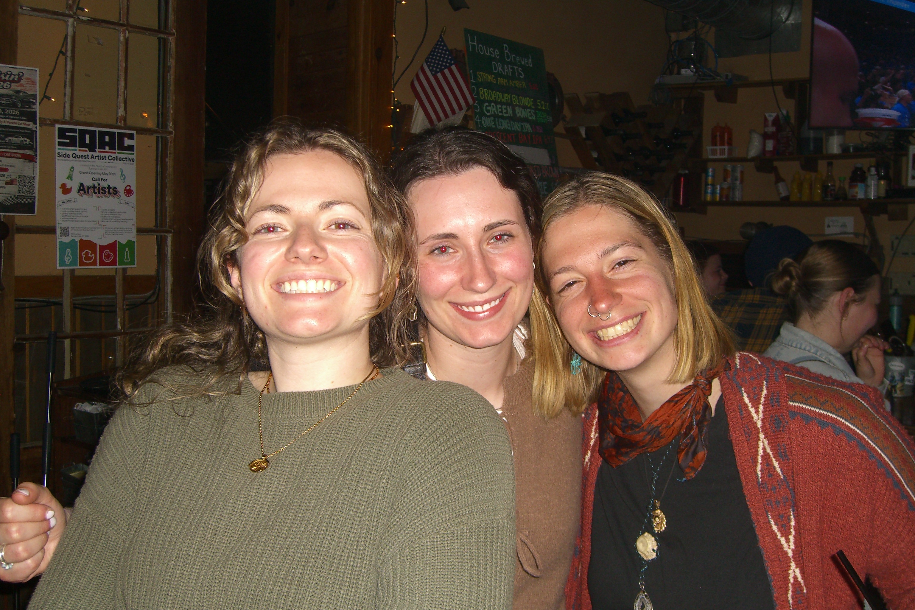 Three women at a bar with a Side Quest Artist Collective poster